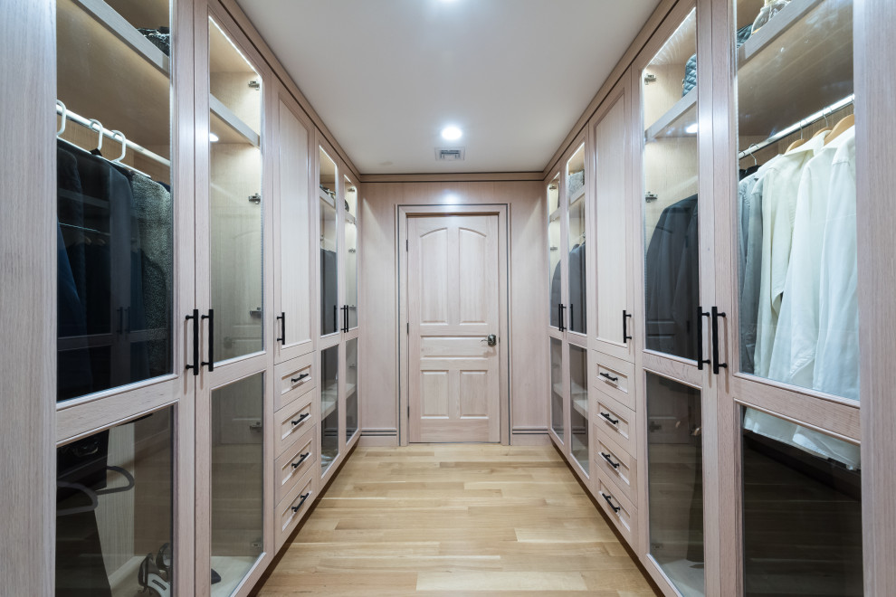 Photo of a mid-sized modern gender-neutral dressing room in New York with glass-front cabinets, light wood cabinets, light hardwood floors and brown floor.