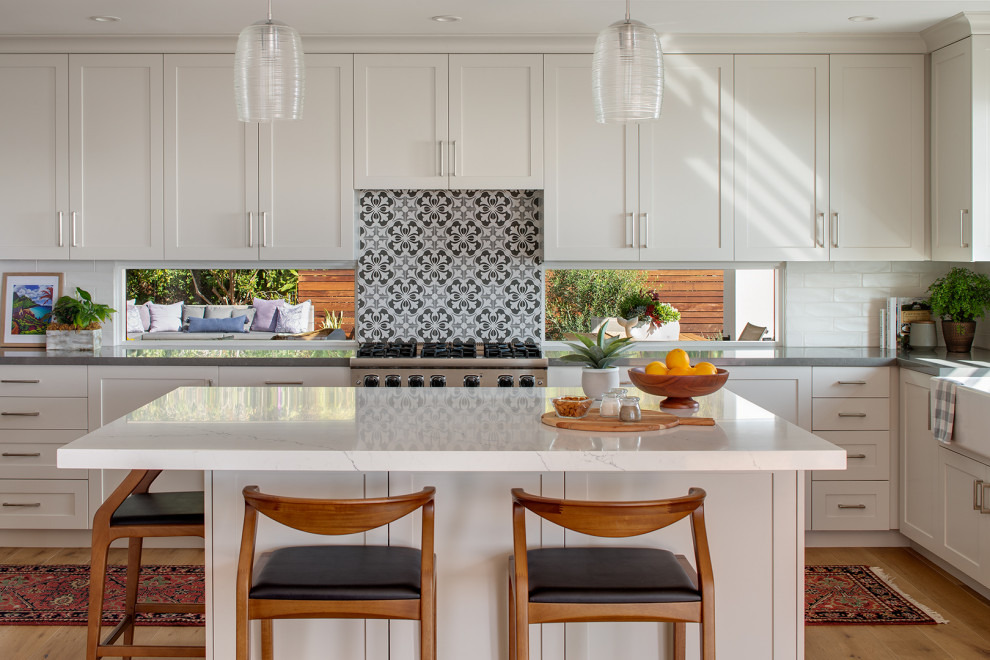 Eat-in kitchen - mid-sized transitional l-shaped light wood floor and brown floor eat-in kitchen idea in San Diego with a farmhouse sink, shaker cabinets, white cabinets, quartz countertops, multicolored backsplash, ceramic backsplash, stainless steel appliances, an island and white countertops