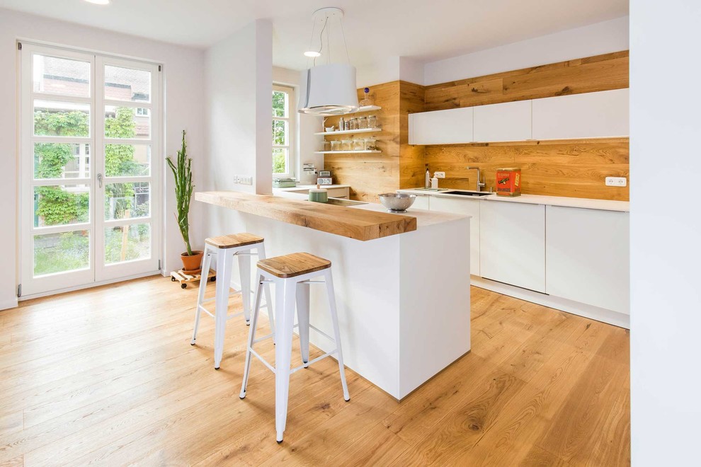 Photo of a mid-sized scandinavian kitchen in Nuremberg with flat-panel cabinets, white cabinets, timber splashback, medium hardwood floors, a peninsula, white benchtop and an undermount sink.