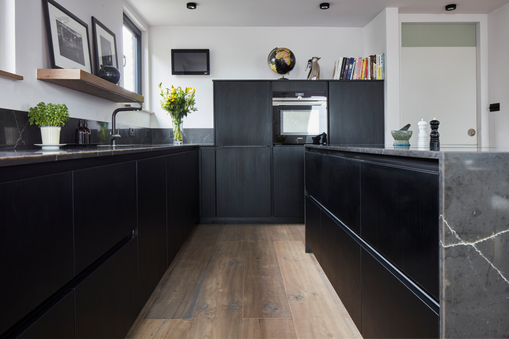 Inredning av ett modernt litet grå grått kök, med en undermonterad diskho, släta luckor, svarta skåp, bänkskiva i kvartsit, grått stänkskydd, rostfria vitvaror, ljust trägolv, en köksö och brunt golv