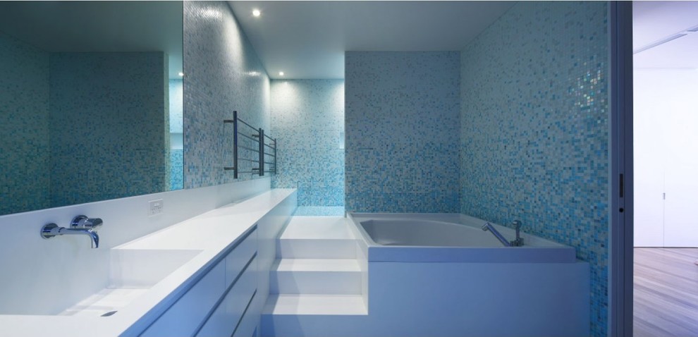 Photo of a large contemporary master bathroom in Brisbane with a drop-in sink, recessed-panel cabinets, white cabinets, solid surface benchtops, a corner tub, white tile and blue walls.