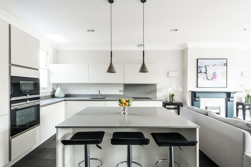 Contemporary l-shaped open plan kitchen in London with flat-panel cabinets, white cabinets, solid surface benchtops, stone tile splashback, stainless steel appliances, dark hardwood floors, with island, a single-bowl sink and grey splashback.