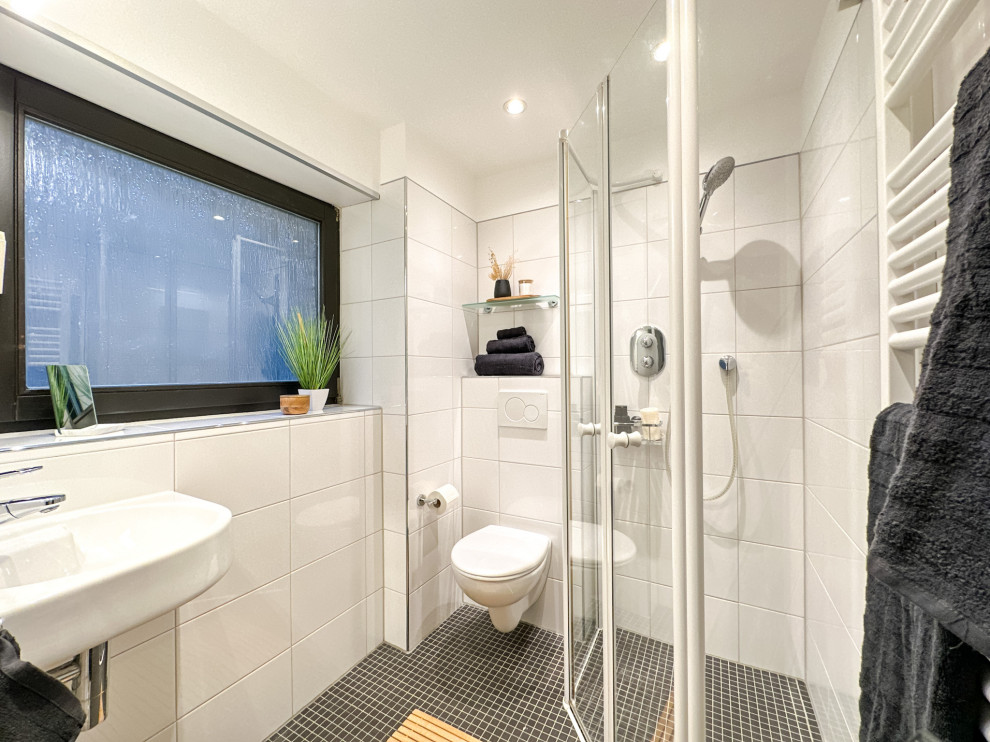 Small contemporary shower room bathroom in Dortmund with a built-in shower, a wall mounted toilet, white tiles, ceramic tiles, white walls, mosaic tile flooring, black floors, a hinged door, a single sink and a built in vanity unit.