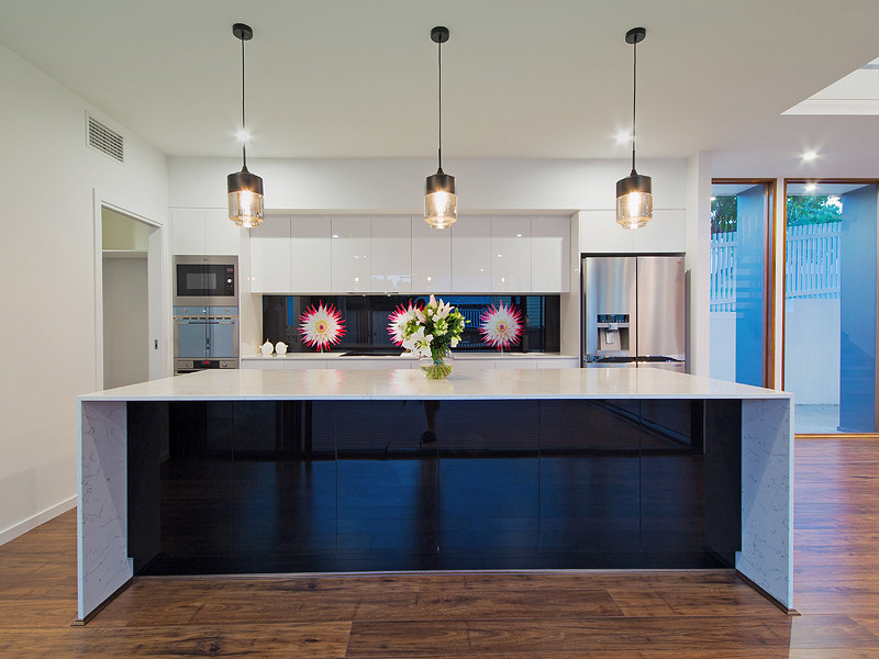 This is an example of a large transitional kitchen in Brisbane with an undermount sink, beaded inset cabinets, white cabinets, granite benchtops, black splashback, glass sheet splashback, stainless steel appliances, medium hardwood floors, with island, brown floor and white benchtop.