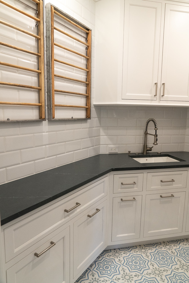 Mid-sized traditional u-shaped dedicated laundry room in DC Metro with an undermount sink, recessed-panel cabinets, white cabinets, soapstone benchtops, vinyl floors, a stacked washer and dryer, multi-coloured floor and grey benchtop.