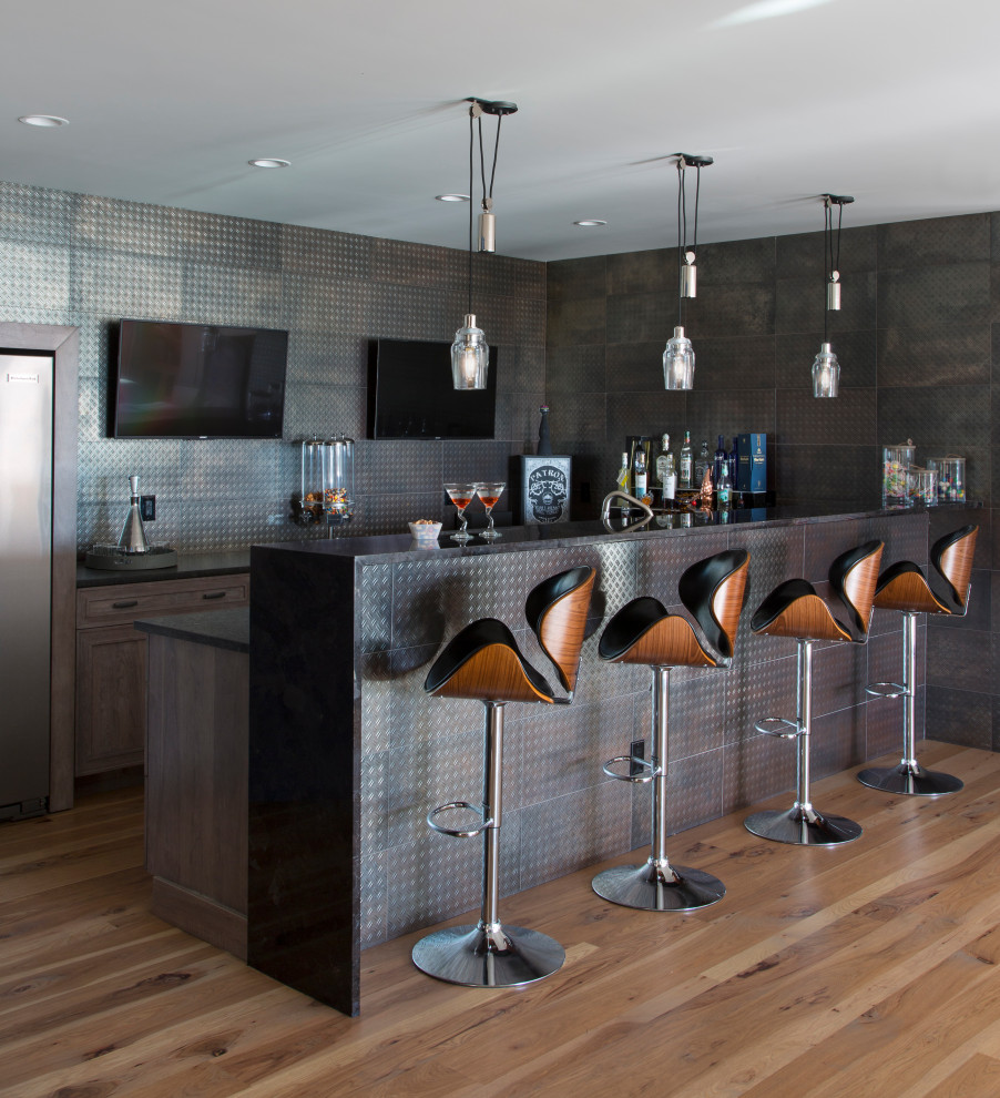 Large contemporary galley seated home bar in Denver with an undermount sink, shaker cabinets, brown cabinets, granite benchtops, multi-coloured splashback, metal splashback, light hardwood floors, brown floor and black benchtop.