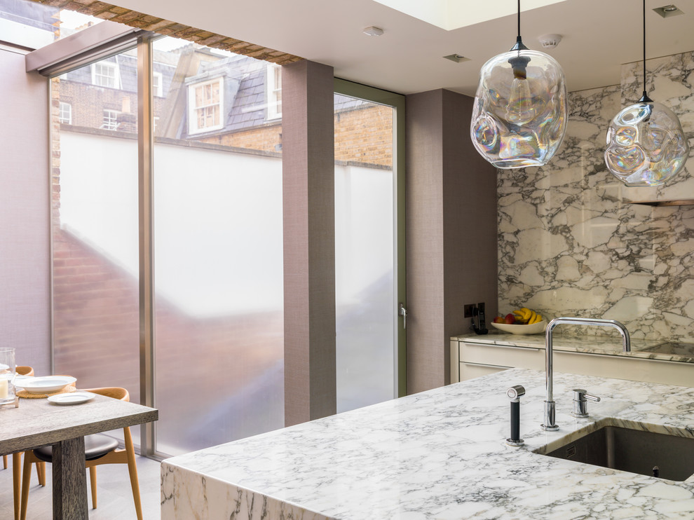 Mid-sized traditional u-shaped open plan kitchen in London with a drop-in sink, louvered cabinets, beige cabinets, marble benchtops, white splashback, marble splashback, limestone floors, with island and beige floor.