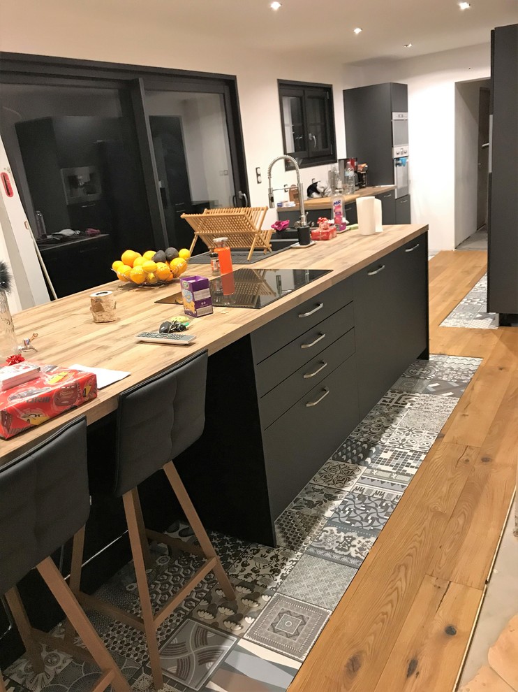 Photo of a large midcentury galley open plan kitchen in Montpellier with ceramic floors, with island, grey floor, a single-bowl sink, flat-panel cabinets, black cabinets, wood benchtops, white splashback, brick splashback, panelled appliances and brown benchtop.
