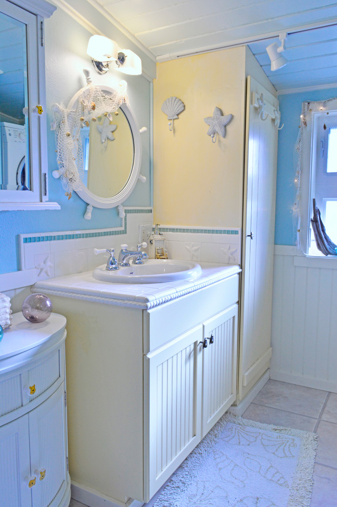 This is an example of a small beach style 3/4 bathroom in Seattle with recessed-panel cabinets, yellow cabinets, white tile, ceramic tile, blue walls, vinyl floors, a drop-in sink, tile benchtops and beige floor.