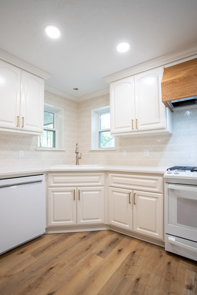 This is an example of a large l-shaped enclosed kitchen in Houston with a built-in sink, raised-panel cabinets, white cabinets, engineered stone countertops, pink splashback, ceramic splashback, white appliances, light hardwood flooring, brown floors and white worktops.