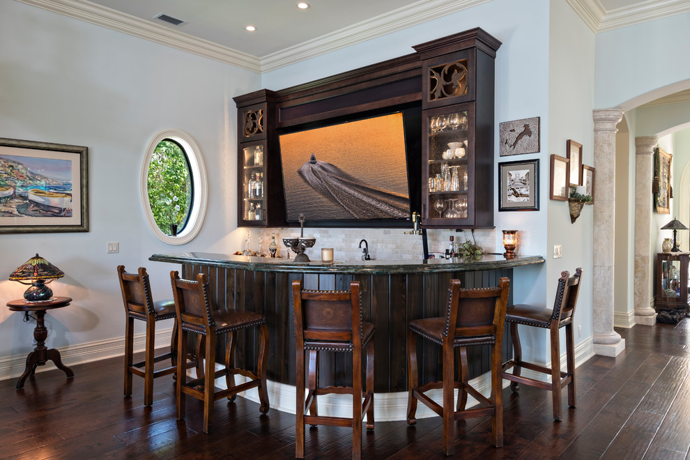 This is an example of a large mediterranean l-shaped seated home bar in Miami with an undermount sink, recessed-panel cabinets, dark wood cabinets, granite benchtops, beige splashback, travertine splashback, dark hardwood floors and brown floor.