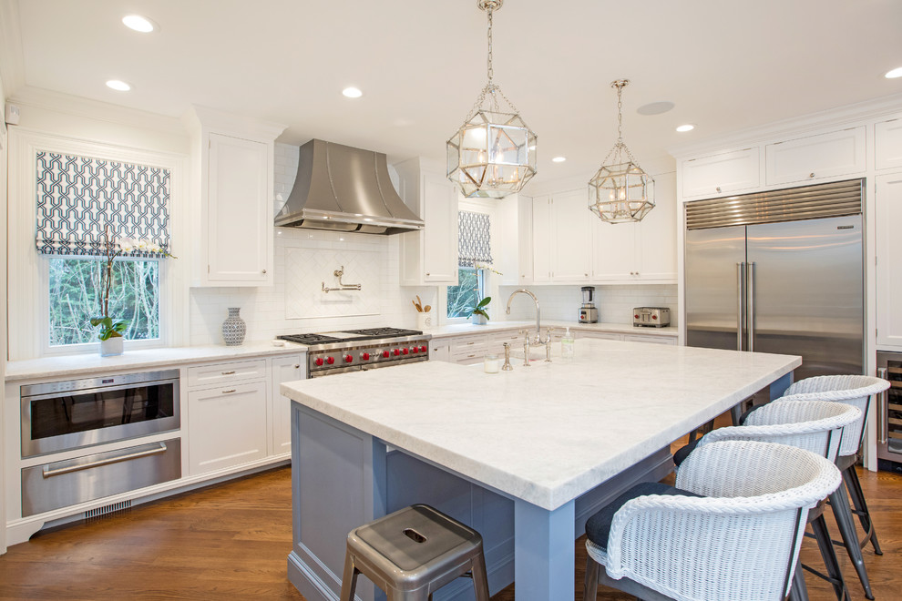 Kitchen - transitional l-shaped medium tone wood floor and brown floor kitchen idea in Miami with a farmhouse sink, recessed-panel cabinets, white cabinets, white backsplash, subway tile backsplash, stainless steel appliances, an island and white countertops