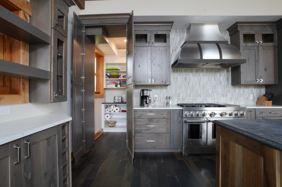 This is an example of a large country l-shaped open plan kitchen in Other with a drop-in sink, flat-panel cabinets, distressed cabinets, quartzite benchtops, grey splashback, ceramic splashback, stainless steel appliances, dark hardwood floors, with island, brown floor and black benchtop.