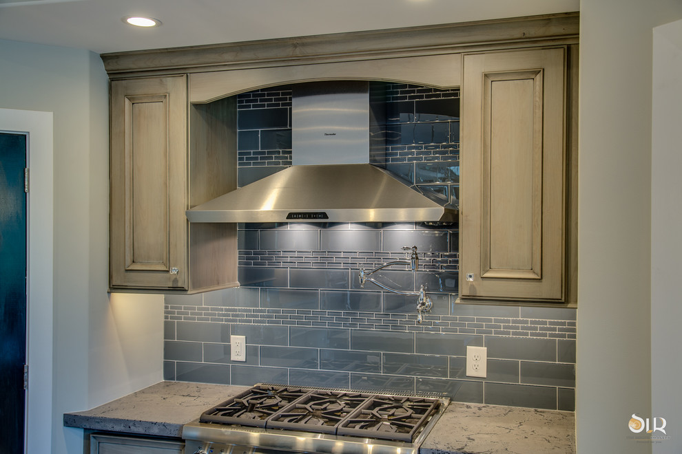 Example of a transitional l-shaped medium tone wood floor and brown floor enclosed kitchen design with an undermount sink, raised-panel cabinets, distressed cabinets, soapstone countertops, gray backsplash, subway tile backsplash, stainless steel appliances and an island