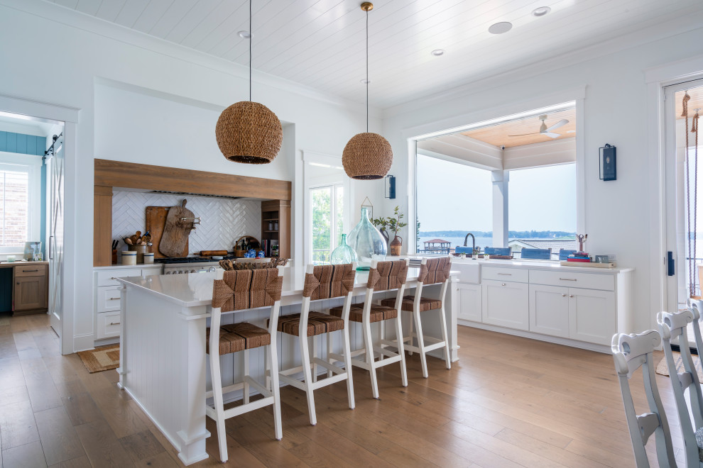 Photo of a large beach style eat-in kitchen in Charlotte with a farmhouse sink, recessed-panel cabinets, white cabinets, marble benchtops, white splashback, stainless steel appliances, brown floor, white benchtop, timber and with island.