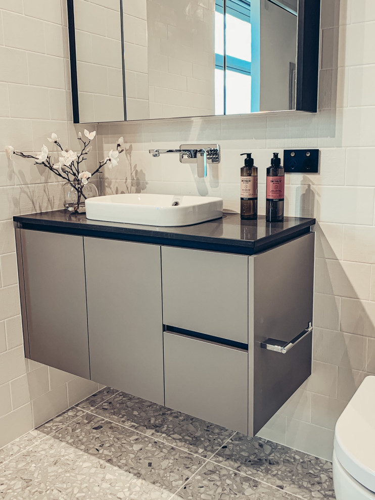 This is an example of a mid-sized contemporary kids bathroom in Sydney with flat-panel cabinets, grey cabinets, a freestanding tub, a corner shower, a two-piece toilet, white tile, ceramic tile, white walls, a vessel sink, engineered quartz benchtops, multi-coloured floor, a hinged shower door, black benchtops, a niche, a single vanity and a floating vanity.