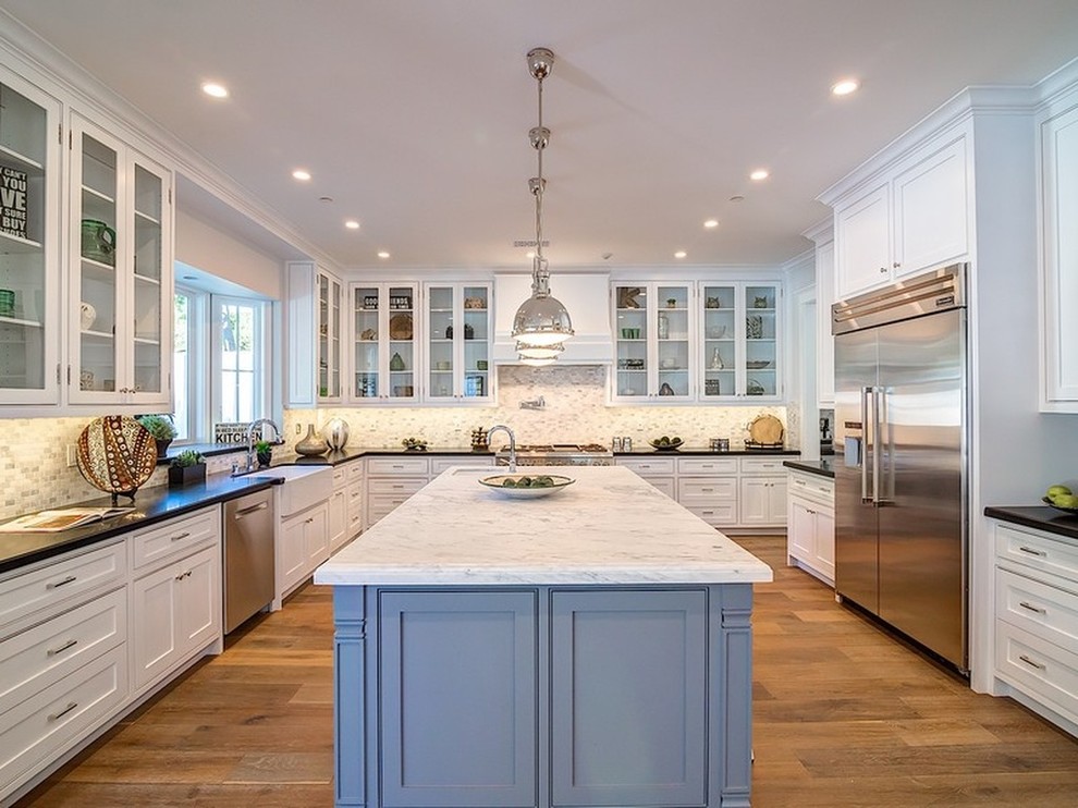 This is an example of an expansive transitional kitchen in Los Angeles with a farmhouse sink, glass-front cabinets, white cabinets, white splashback, subway tile splashback, stainless steel appliances, medium hardwood floors and with island.