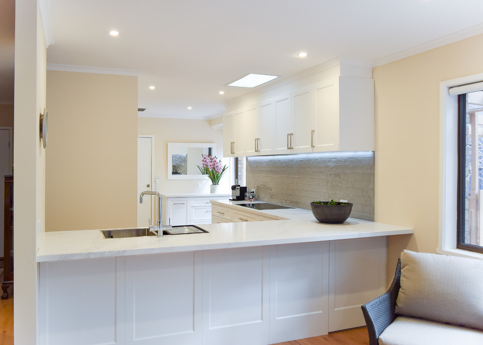 This is an example of a mid-sized transitional l-shaped kitchen pantry in Melbourne with an undermount sink, shaker cabinets, white cabinets, quartz benchtops, beige splashback, subway tile splashback, stainless steel appliances, light hardwood floors, no island and brown floor.