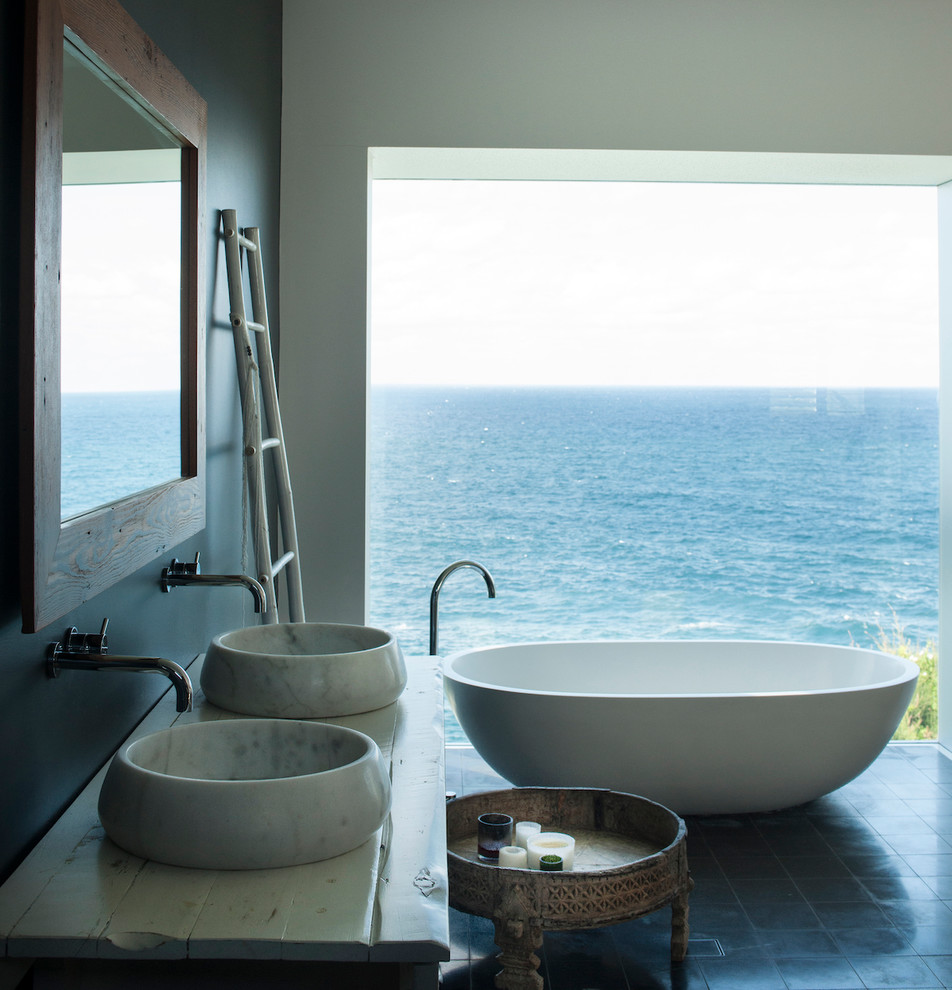 Photo of a beach style master bathroom in Sydney with beige cabinets, a freestanding tub, black walls, a vessel sink, wood benchtops, black floor and white benchtops.