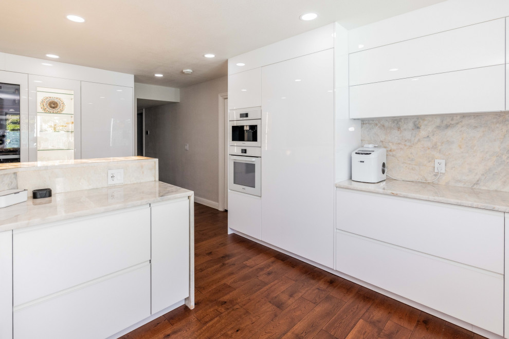Photo of a mid-sized contemporary u-shaped eat-in kitchen in San Francisco with an integrated sink, flat-panel cabinets, white cabinets, quartz benchtops, beige splashback, engineered quartz splashback, panelled appliances, cork floors, with island, brown floor and black benchtop.