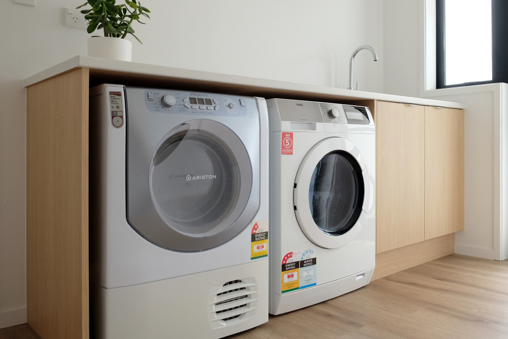 Photo of a large contemporary laundry room in Other with an undermount sink, flat-panel cabinets, light wood cabinets, solid surface benchtops, white splashback, ceramic splashback, laminate floors, brown floor and white benchtop.