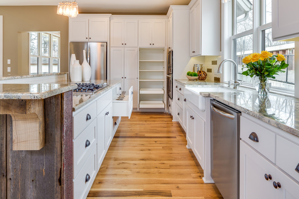 Mid-sized arts and crafts l-shaped eat-in kitchen in Other with a farmhouse sink, recessed-panel cabinets, white cabinets, granite benchtops, white splashback, ceramic splashback, stainless steel appliances, light hardwood floors, with island and beige floor.