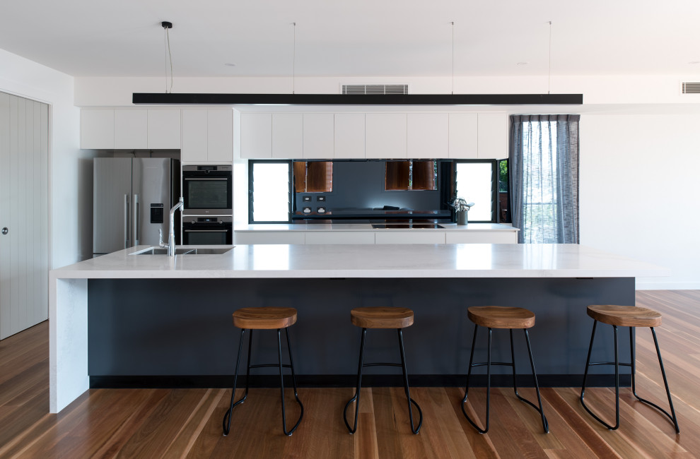 This is an example of a mid-sized contemporary galley open plan kitchen in Brisbane with an undermount sink, flat-panel cabinets, white cabinets, quartz benchtops, white splashback, glass sheet splashback, black appliances, with island, brown floor, white benchtop and medium hardwood floors.