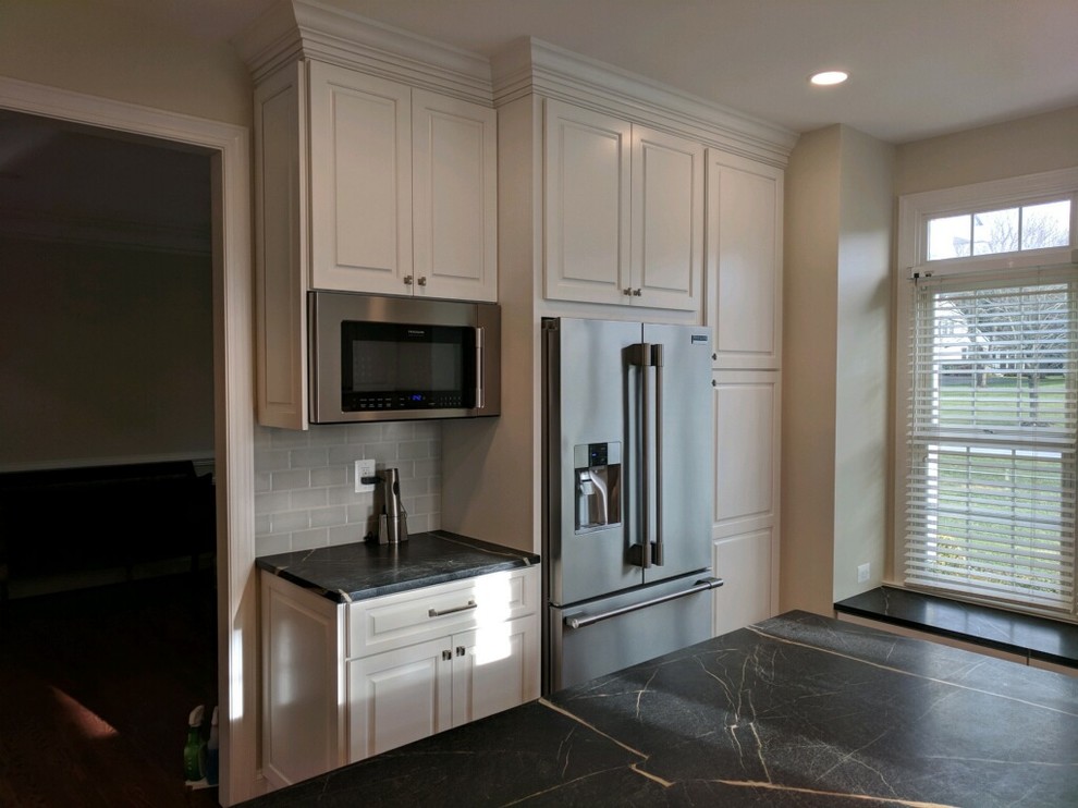 Large transitional l-shaped dark wood floor and brown floor eat-in kitchen photo in Baltimore with a farmhouse sink, raised-panel cabinets, white cabinets, soapstone countertops, gray backsplash, subway tile backsplash, stainless steel appliances and an island