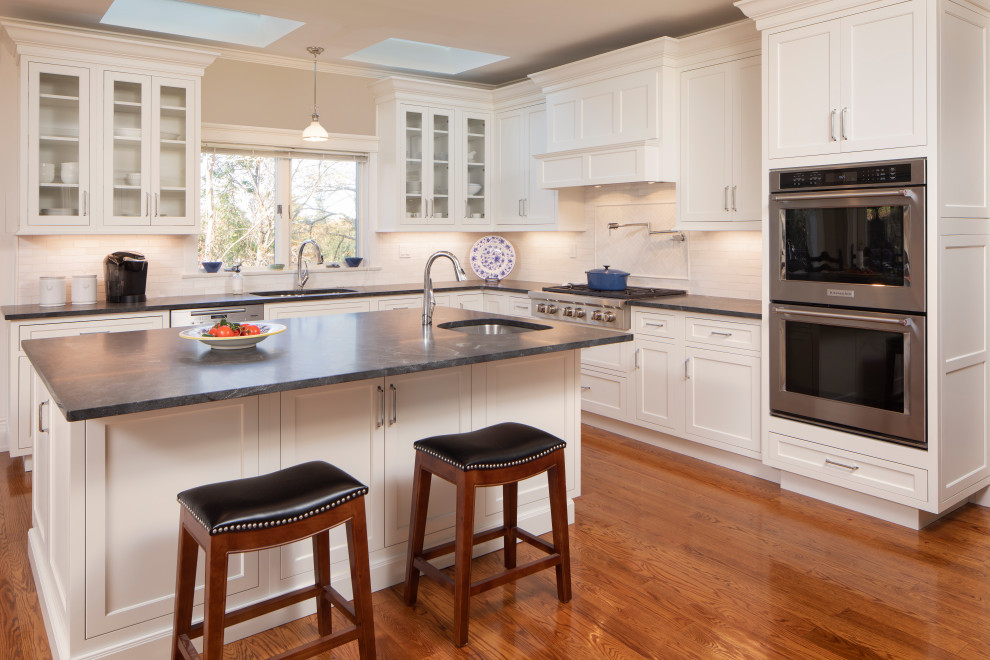 Large traditional l-shaped eat-in kitchen in New York with an undermount sink, shaker cabinets, white cabinets, granite benchtops, white splashback, subway tile splashback, stainless steel appliances, medium hardwood floors, with island, beige floor, black benchtop and exposed beam.