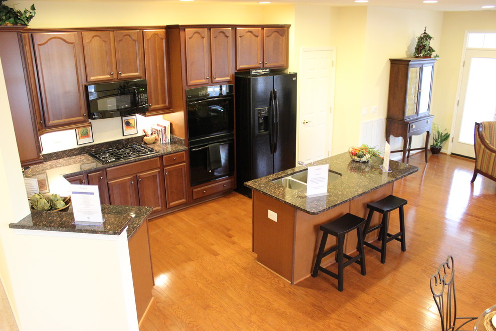 Example of a l-shaped medium tone wood floor eat-in kitchen design with dark wood cabinets, granite countertops, beige backsplash, black appliances and an island