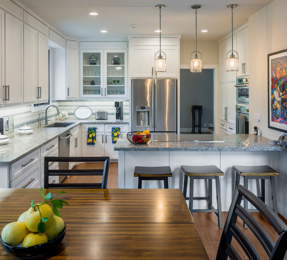 Example of a transitional l-shaped medium tone wood floor and brown floor kitchen design in Houston with an undermount sink, recessed-panel cabinets, white cabinets, gray backsplash, mosaic tile backsplash, stainless steel appliances, a peninsula and gray countertops