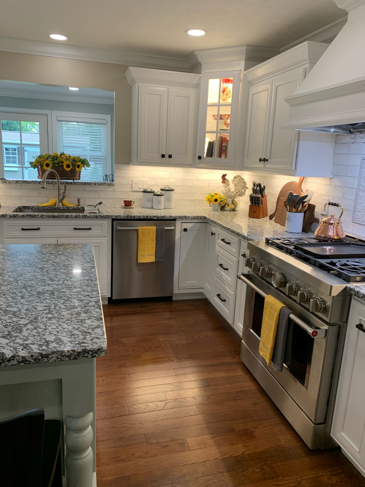 Example of a transitional l-shaped medium tone wood floor and brown floor kitchen design with an undermount sink, recessed-panel cabinets, white cabinets, granite countertops, ceramic backsplash, stainless steel appliances, an island and gray countertops