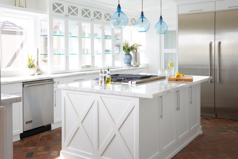 Inspiration for a mid-sized beach style l-shaped separate kitchen in Los Angeles with recessed-panel cabinets, white cabinets, stainless steel appliances, brick floors, with island, marble benchtops, window splashback and red floor.