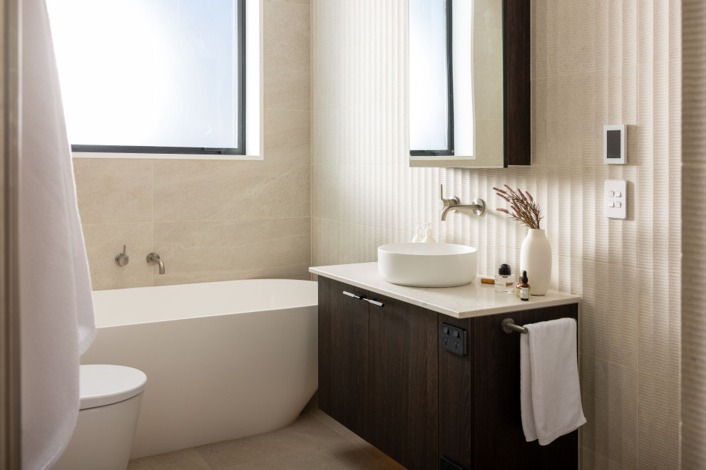 Стильный дизайн: главная ванная комната среднего размера в стиле модернизм с коричневыми фасадами, отдельно стоящей ванной, открытым душем, унитазом-моноблоком, бежевой плиткой, каменной плиткой, бежевыми стенами, настольной раковиной, столешницей из дерева, открытым душем, белой столешницей, тумбой под одну раковину и подвесной тумбой - последний тренд
