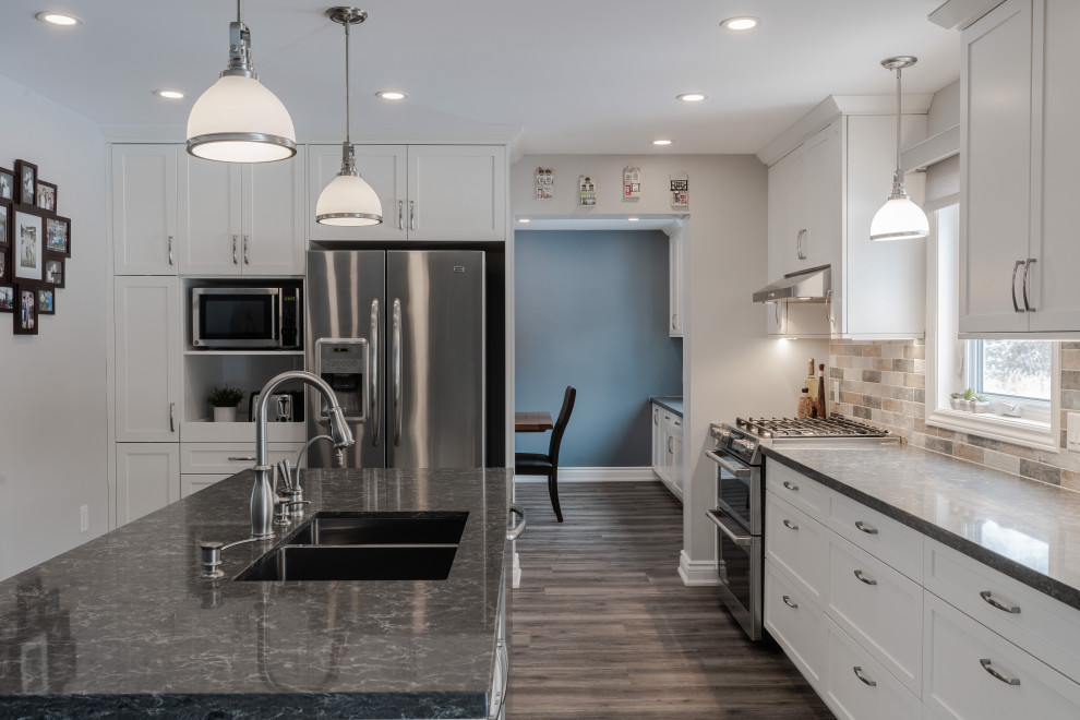 Design ideas for a mid-sized country l-shaped kitchen in Toronto with a double-bowl sink, shaker cabinets, white cabinets, quartzite benchtops, multi-coloured splashback, brick splashback, stainless steel appliances, vinyl floors, with island, brown floor and grey benchtop.