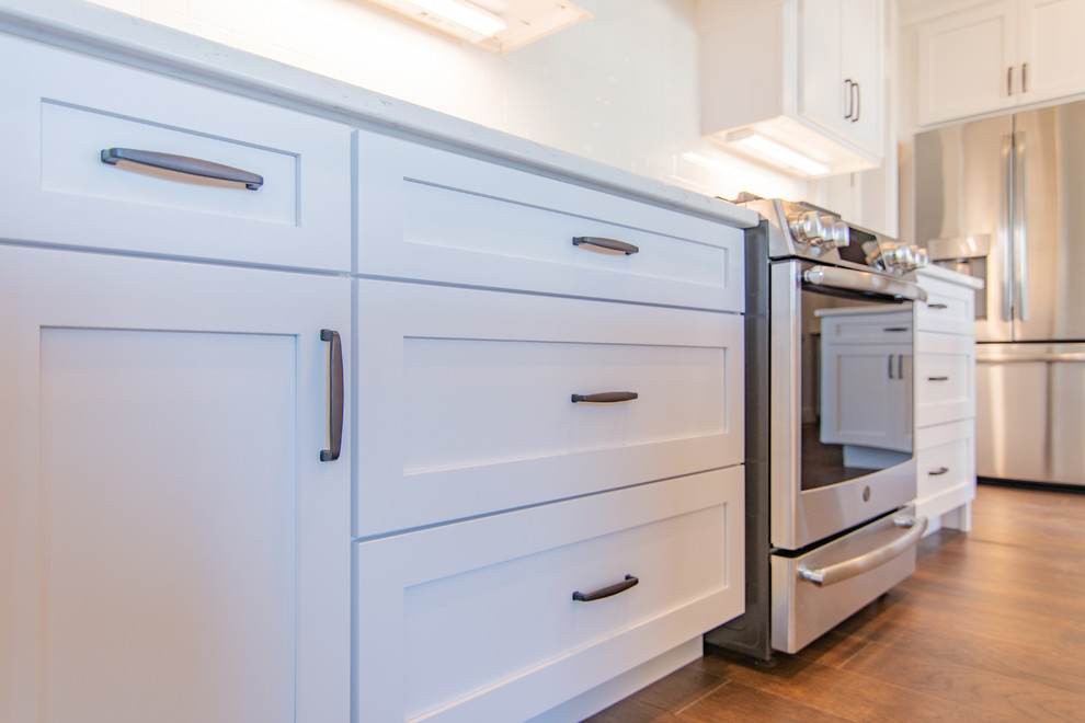 Transitional medium tone wood floor and brown floor kitchen pantry photo with an undermount sink, shaker cabinets, white cabinets, quartz countertops, white backsplash, subway tile backsplash, stainless steel appliances, an island and white countertops