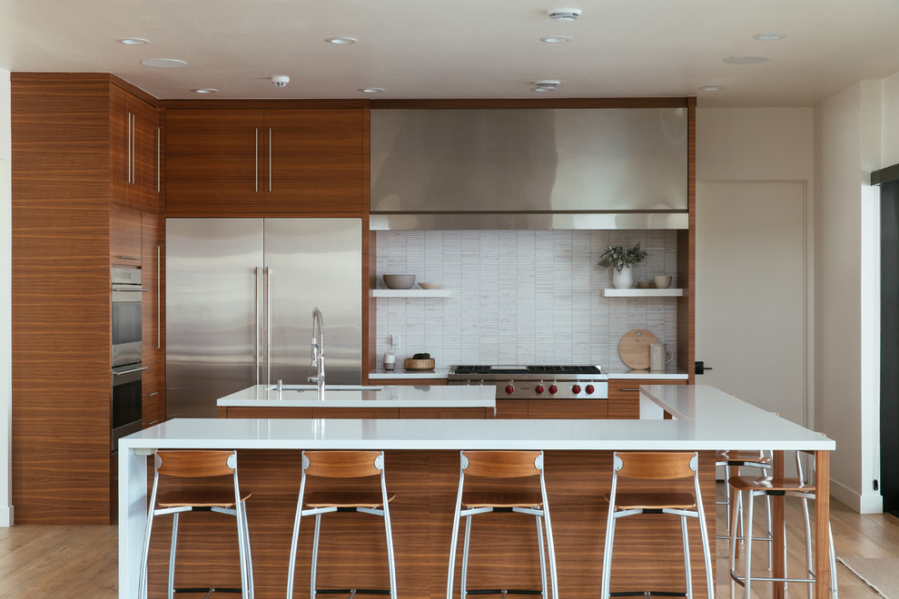 Inspiration for a contemporary l-shaped kitchen in Salt Lake City with an undermount sink, flat-panel cabinets, medium wood cabinets, white splashback, stainless steel appliances, medium hardwood floors, multiple islands, brown floor and white benchtop.