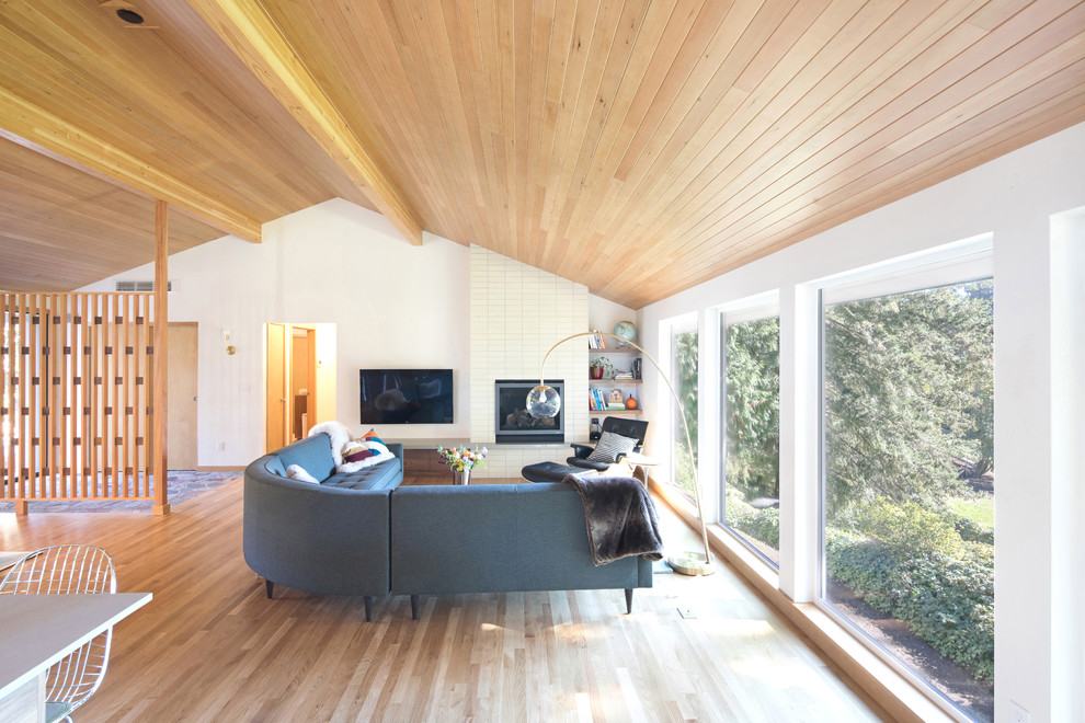 Photo of a mid-sized midcentury open concept family room in Seattle with medium hardwood floors, a standard fireplace, a tile fireplace surround, a wall-mounted tv, brown floor and white walls.