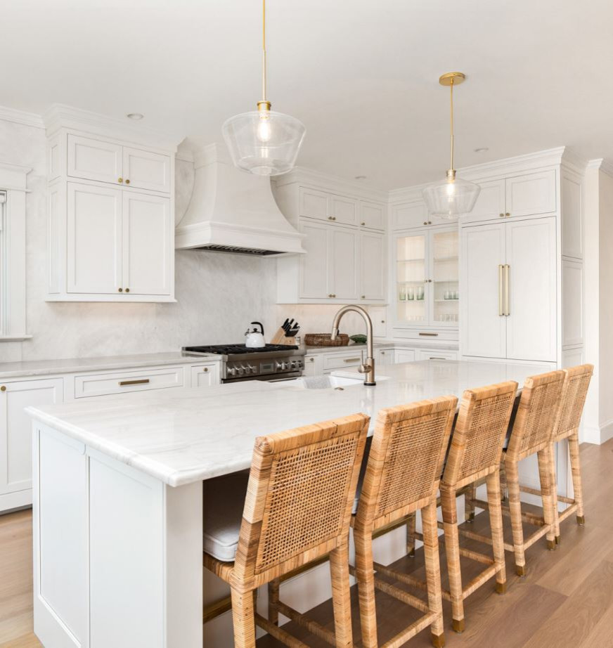 Photo of a large beach style l-shaped kitchen in Portland Maine with a farmhouse sink, shaker cabinets, white cabinets, marble benchtops, white splashback, marble splashback, white appliances, medium hardwood floors, with island, brown floor and white benchtop.