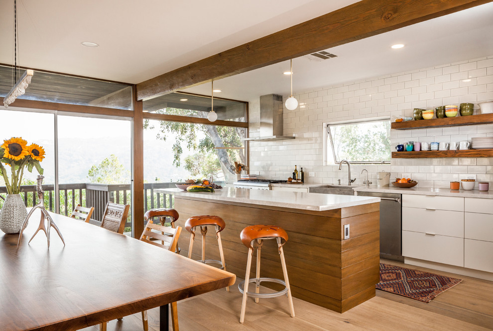 Mid-sized transitional single-wall light wood floor eat-in kitchen photo in Los Angeles with a farmhouse sink, flat-panel cabinets, white cabinets, white backsplash, subway tile backsplash, stainless steel appliances and an island