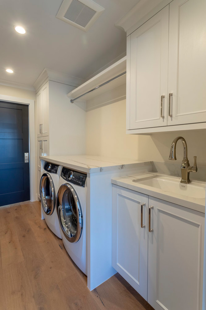 Expansive transitional galley dedicated laundry room in Los Angeles with an undermount sink, recessed-panel cabinets, white cabinets, quartz benchtops, white walls, medium hardwood floors, a side-by-side washer and dryer and white benchtop.