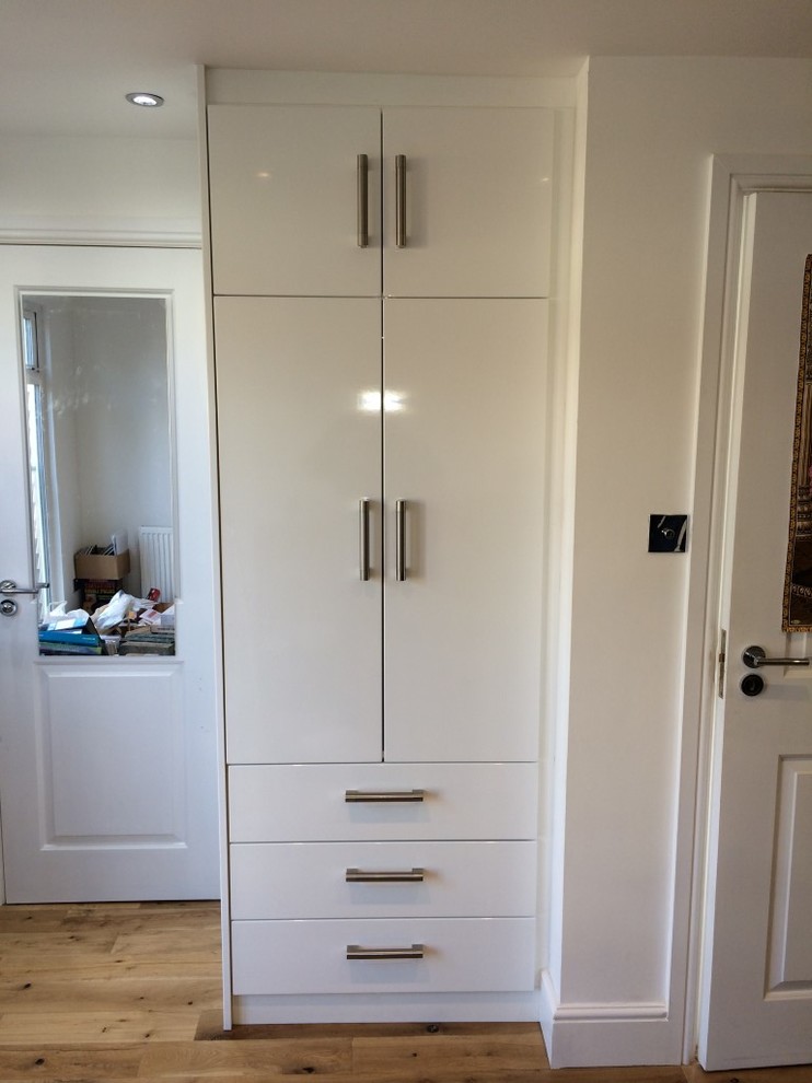 Photo of a small modern gender-neutral built-in wardrobe in London with flat-panel cabinets, white cabinets and light hardwood floors.