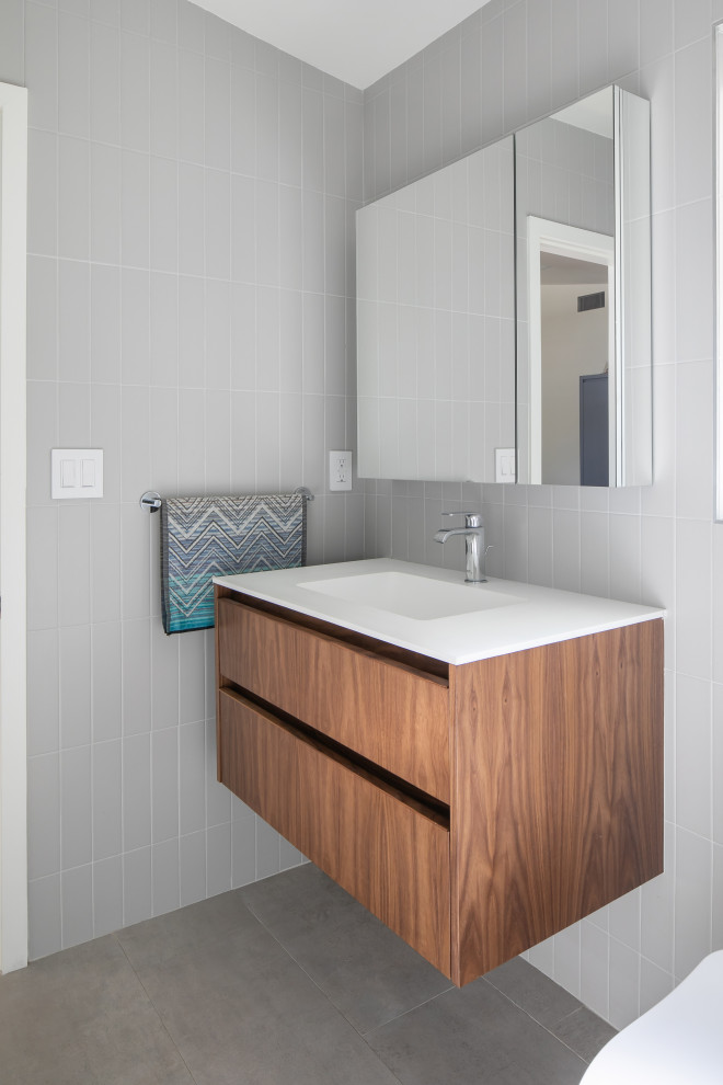 Small retro bathroom in Los Angeles with flat-panel cabinets, medium wood cabinets, an alcove shower, a one-piece toilet, grey tiles, ceramic tiles, grey walls, porcelain flooring, a wall-mounted sink, engineered stone worktops, grey floors, a hinged door, white worktops, a single sink and a freestanding vanity unit.
