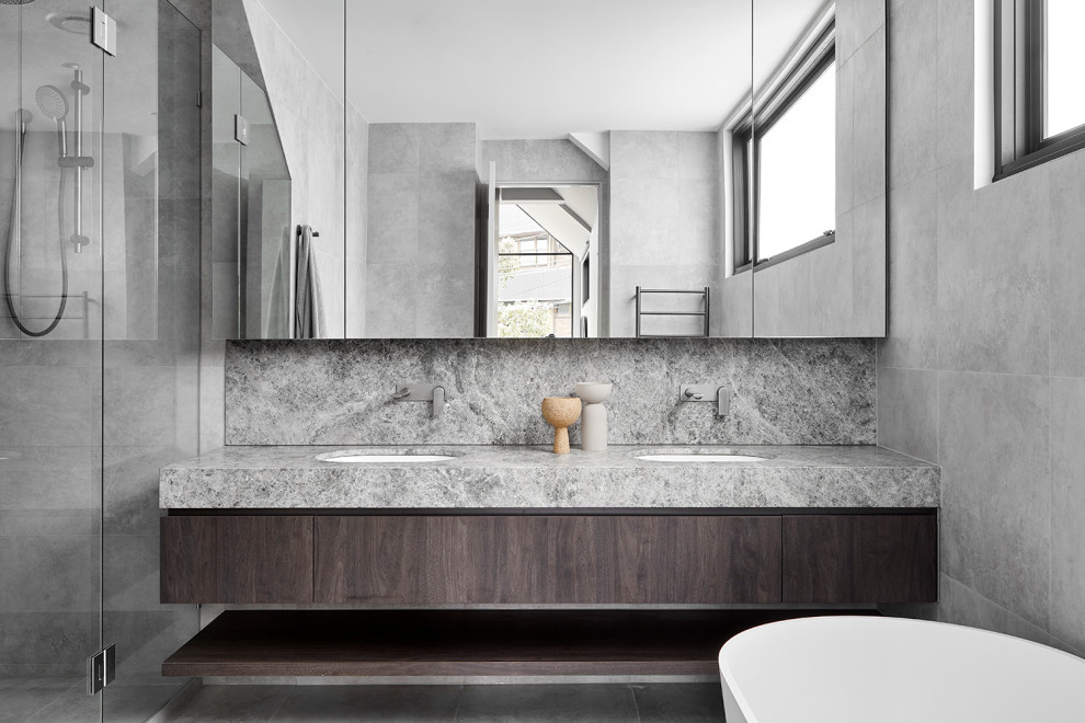 Photo of a mid-sized modern master bathroom in Melbourne with furniture-like cabinets, dark wood cabinets, a freestanding tub, an alcove shower, a one-piece toilet, gray tile, ceramic tile, grey walls, ceramic floors, an integrated sink, limestone benchtops, grey floor, a hinged shower door, grey benchtops, a niche, a double vanity and a built-in vanity.