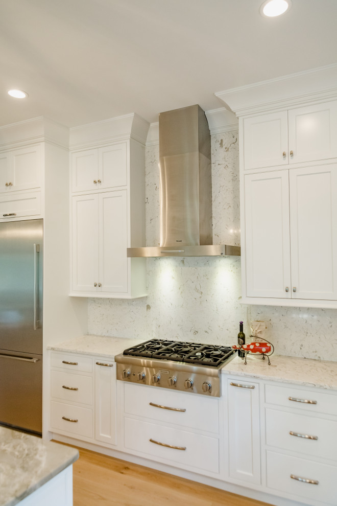 This is an example of a large transitional l-shaped open plan kitchen in Charleston with an undermount sink, beaded inset cabinets, white cabinets, quartz benchtops, white splashback, engineered quartz splashback, stainless steel appliances, light hardwood floors, with island, brown floor and white benchtop.