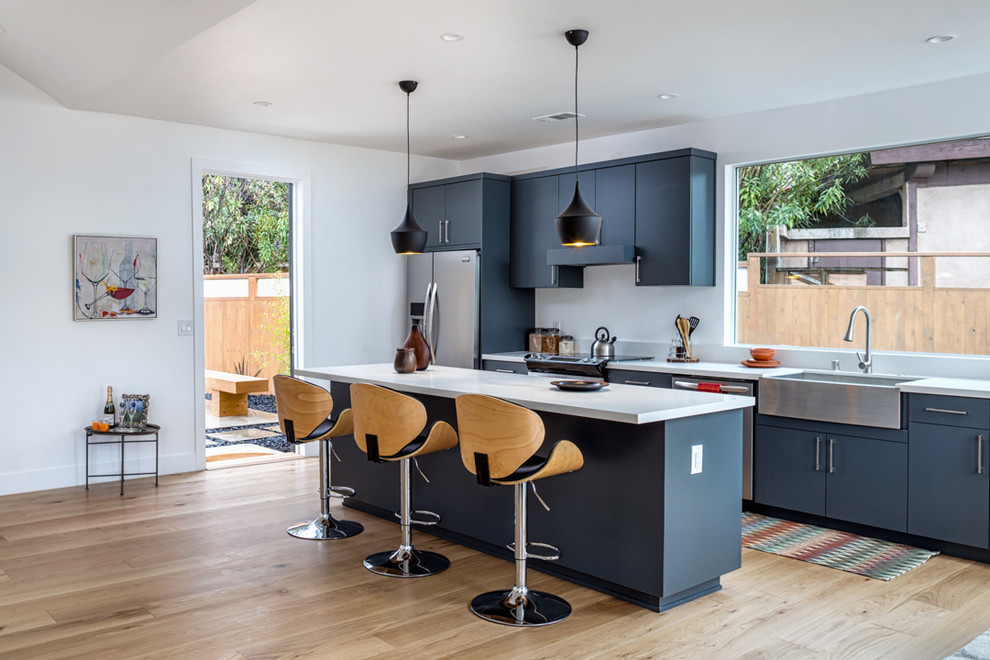 Photo of a contemporary kitchen in Los Angeles with a farmhouse sink, flat-panel cabinets, blue cabinets, window splashback, stainless steel appliances, light hardwood floors and with island.