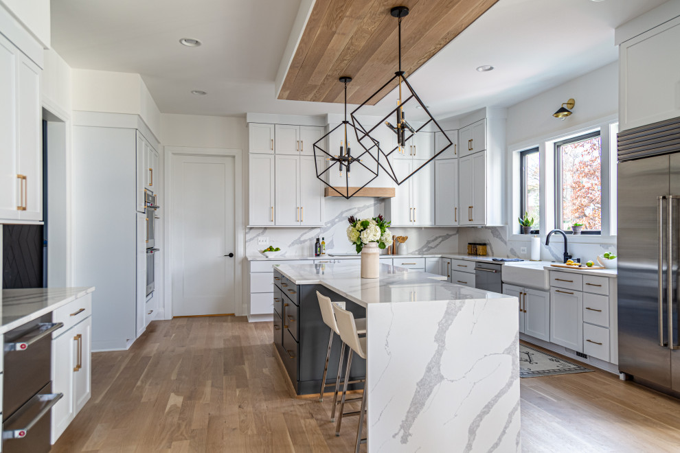 Photo of a large country u-shaped open plan kitchen in DC Metro with a farmhouse sink, shaker cabinets, white cabinets, quartz benchtops, white splashback, engineered quartz splashback, stainless steel appliances, light hardwood floors, with island, beige floor, white benchtop and wood.