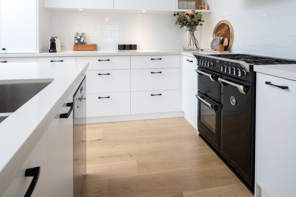 Photo of a modern kitchen in Perth with flat-panel cabinets, white cabinets, quartz benchtops, white splashback, subway tile splashback, black appliances, medium hardwood floors and white benchtop.