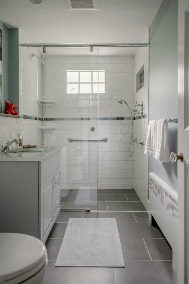 This is an example of a mid-sized transitional master bathroom in Boston with raised-panel cabinets, white cabinets, a curbless shower, a two-piece toilet, white tile, subway tile, grey walls, porcelain floors, an undermount sink, marble benchtops, grey floor, a sliding shower screen and white benchtops.