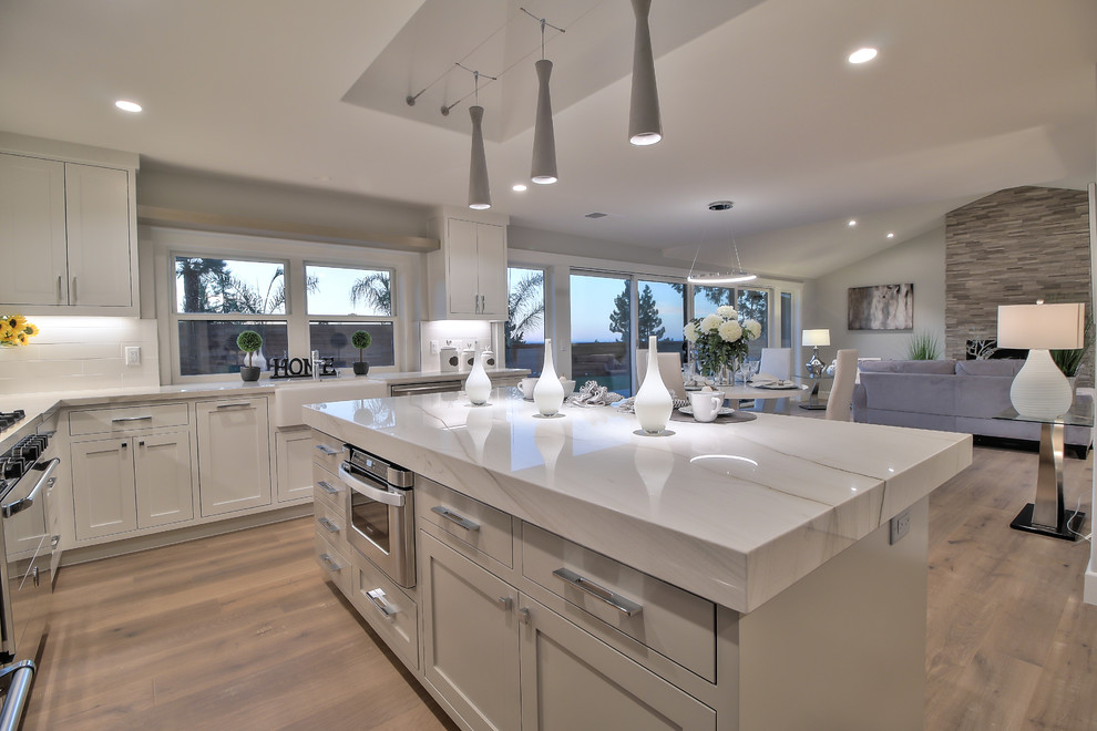 This is an example of a modern l-shaped eat-in kitchen in San Francisco with a farmhouse sink, shaker cabinets, white cabinets, quartzite benchtops, white splashback, porcelain splashback, stainless steel appliances, light hardwood floors, with island and green floor.